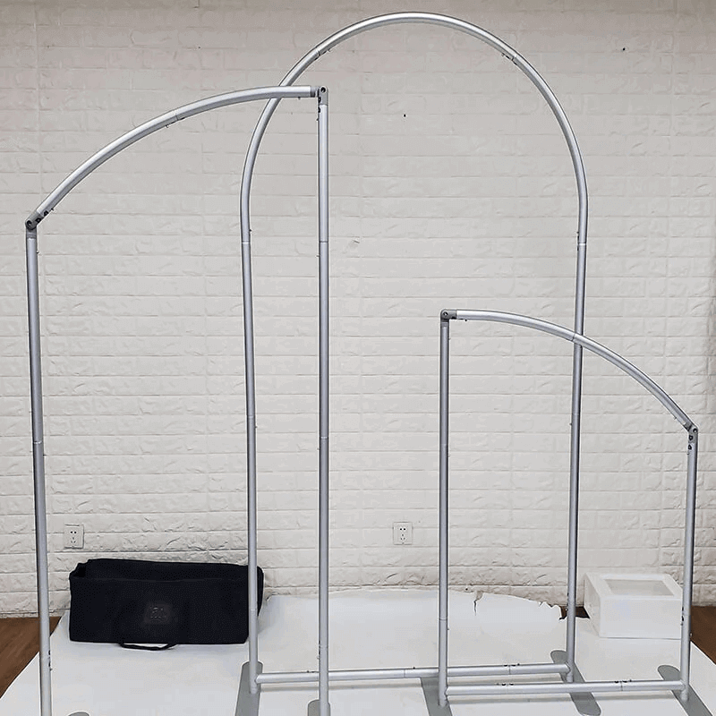 A photo of two metal modular arch frames of different heights. The larger frame has a tall and broad arch, while the smaller frame has a shorter and narrower arch. Both frames are constructed with silver-colored metal tubing and supported by sturdy rectangular bases. A black carrying bag is visible in the background, and the setup is placed on a white surface against a wall with a light brick-like pattern.