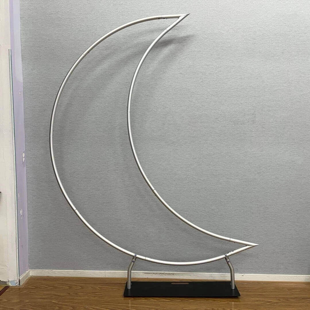 A crescent moon-shaped metal backdrop stand with a sturdy base. The frame is minimalistic, consisting of thin metallic tubing, and is placed against a neutral gray wall on a wooden floor.