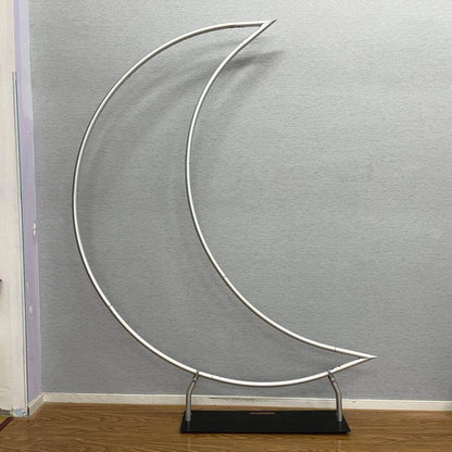 A crescent moon-shaped metal backdrop stand with a sturdy base. The frame is minimalistic, consisting of thin metallic tubing, and is placed against a neutral gray wall on a wooden floor.