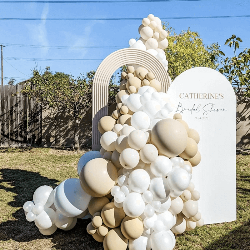 A decorative balloon arrangement featuring white and beige balloons of varying sizes, arranged around a groovy open arch in a backyard setting. The arch is paired with a custom sign that reads "Catherine's Bridal Shower 9.24.2022" on a white background.