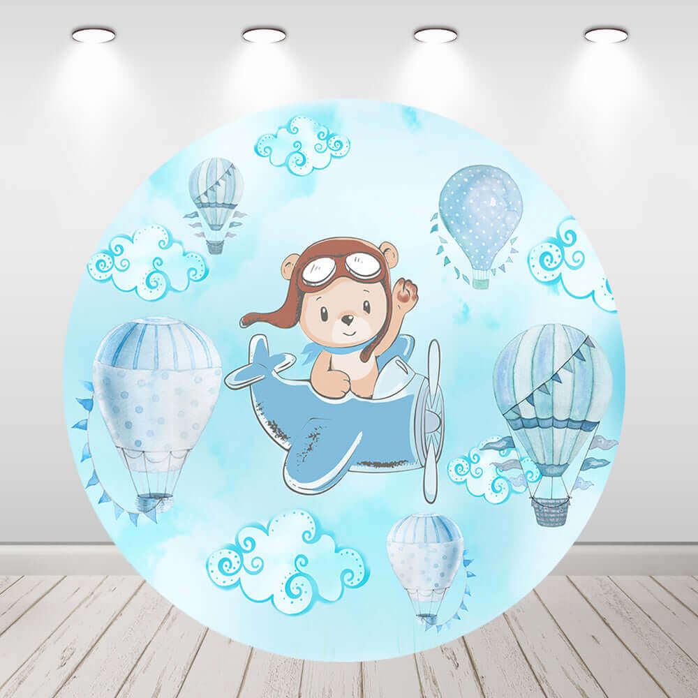 Round backdrop featuring a cartoon pilot bear flying a blue airplane, surrounded by pastel hot air balloons, fluffy clouds, and a light blue sky background, ideal for baby showers and kids’ parties.