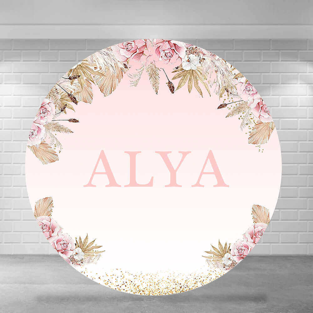 Elegant round backdrop with a blush pink gradient background, personalized with the name "ALYA" in large letters, surrounded by floral arrangements featuring pink roses, gold leaves, and glitter accents.