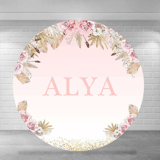 Elegant round backdrop with a blush pink gradient background, personalized with the name "ALYA" in large letters, surrounded by floral arrangements featuring pink roses, gold leaves, and glitter accents.