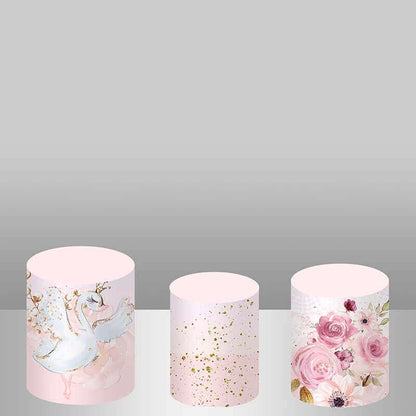 Set of three plinth covers for swan-themed party decor: one with swans and florals, one with gold splatter on pink, and one with lush pink floral arrangements.