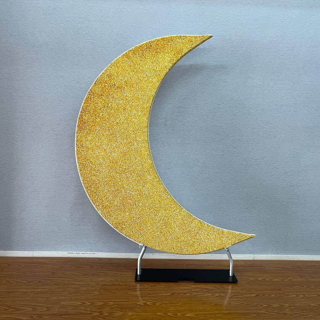 Sparkling gold crescent moon-shaped backdrop stand on a wooden floor against a neutral wall, ideal for party decor, weddings, and photo booths.