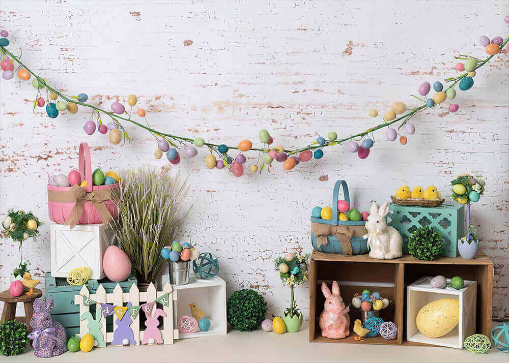 Spring Easter-themed backdrop with a white brick wall, pastel egg garlands, bunny figurines, decorative baskets, greenery, and wooden crates, perfect for seasonal celebrations and photos.