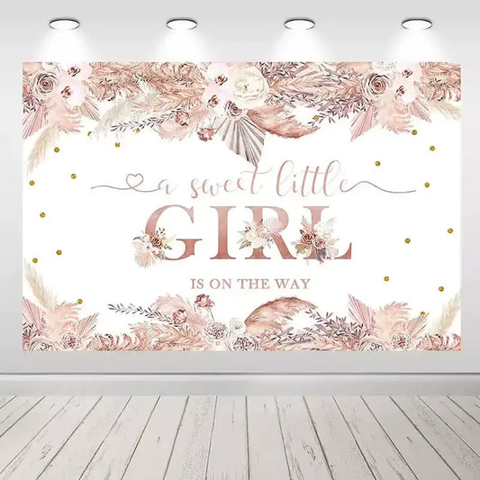 A pink boho-themed baby shower backdrop with floral arrangements, pampas grass, gold accents, and the phrase 'A Sweet Little Girl Is On The Way' in elegant text.