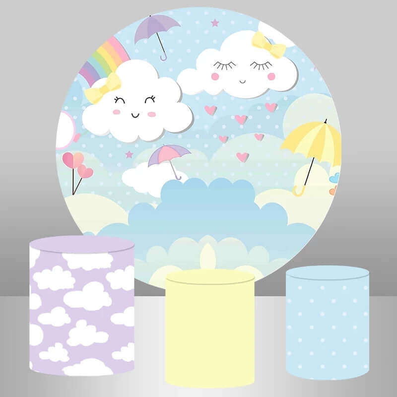 Pastel-themed party backdrop featuring smiling clouds, a rainbow, heart-shaped balloons, and umbrellas, paired with plinth covers in purple with white clouds, solid pastel yellow, and blue polka dots.