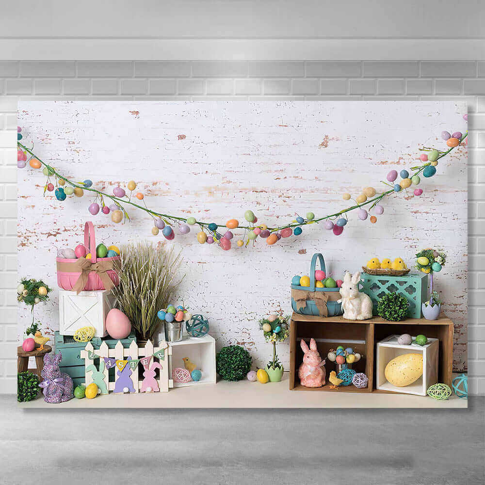 Spring Easter-themed backdrop with a white brick wall, pastel egg garlands, bunny figurines, decorative baskets, greenery, and wooden crates, perfect for seasonal celebrations and photos.